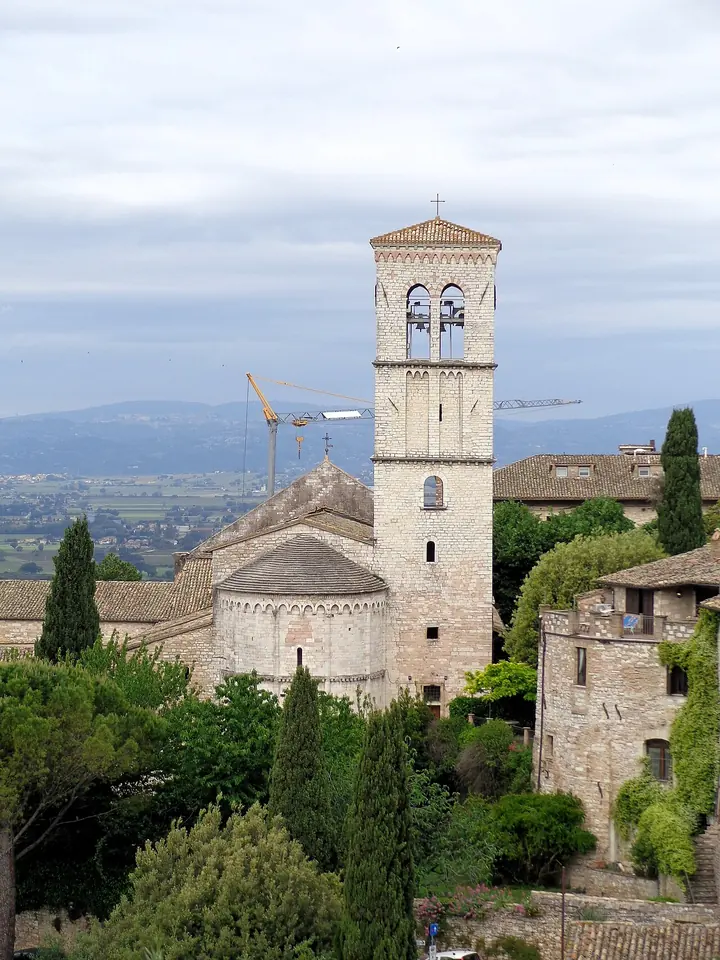 Ábside redondeado de piedra y alto campanario cuadrado de una iglesia románica entre cipreses y tejados, con la llanura de Umbría al fondo
