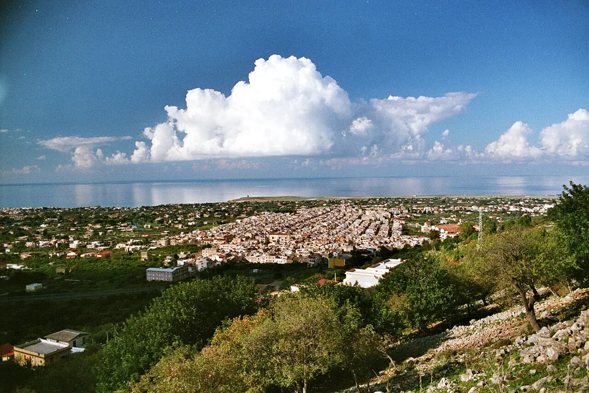The town of Cinisi spread across a green coastal plain below wooded hills, with the blue sea and a towering white cloud beyond