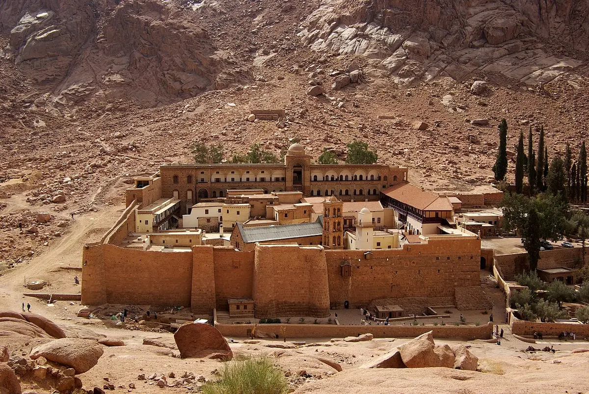 The fortified stone walls and buildings of St. Catherine's Monastery at the foot of bare granite mountains in the Sinai desert, with a few cypress trees