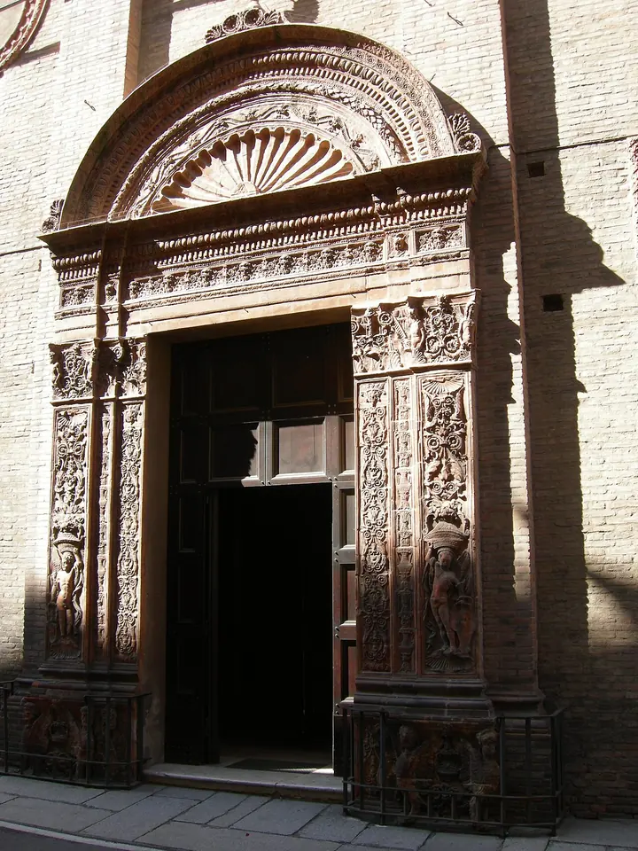 A richly carved Renaissance terracotta doorway with a shell-shaped arch, set in a plain brick church front on a narrow Bologna street