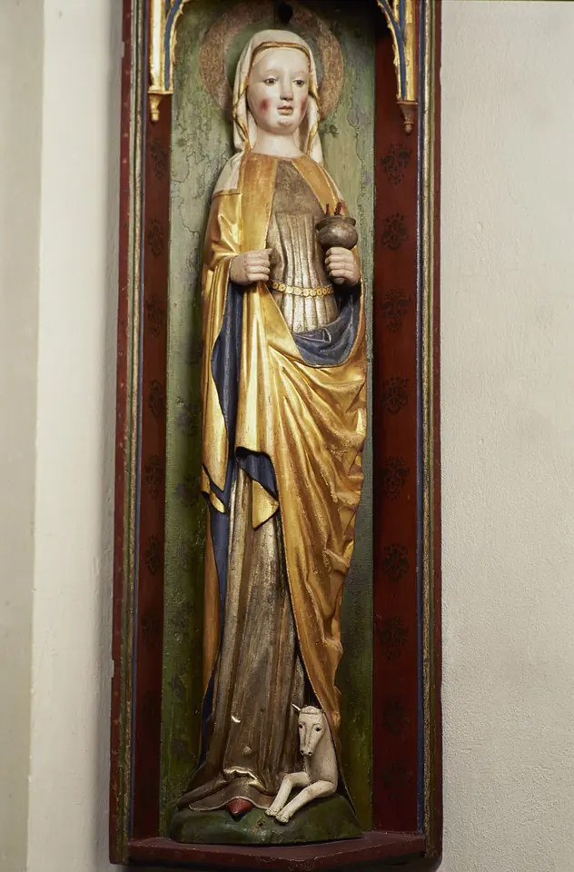 A late medieval gilded wooden statue of a veiled woman in a long gold mantle standing in a red niche, with a small hind lying at her feet