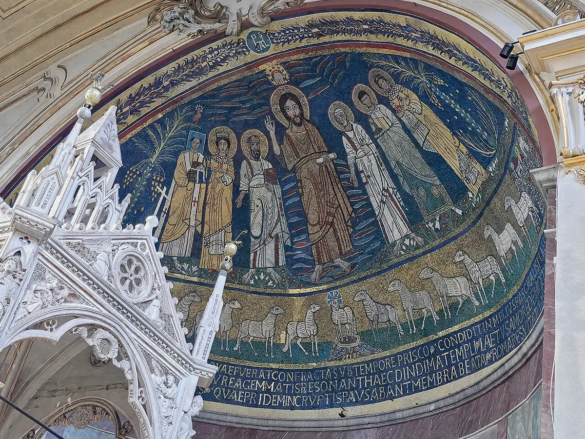 Mosaico dorado y azul del ábside con Cristo de pie entre santos y una hilera de corderos debajo, visto sobre los pináculos góticos blancos del baldaquino de mármol