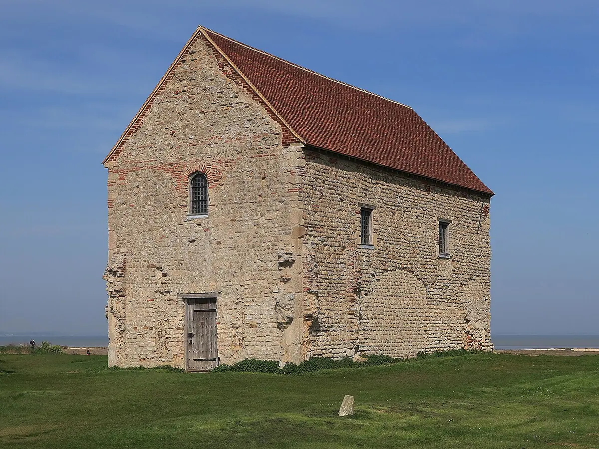 A tall, plain stone chapel with a steep red-tiled roof standing alone on green grass at the edge of the sea under a blue sky