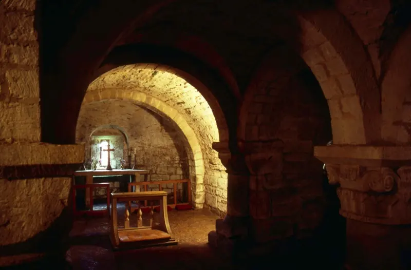 Low vaulted stone crypt with thick round columns and carved capitals, lit warmly, with a small altar and a cross beneath a little window
