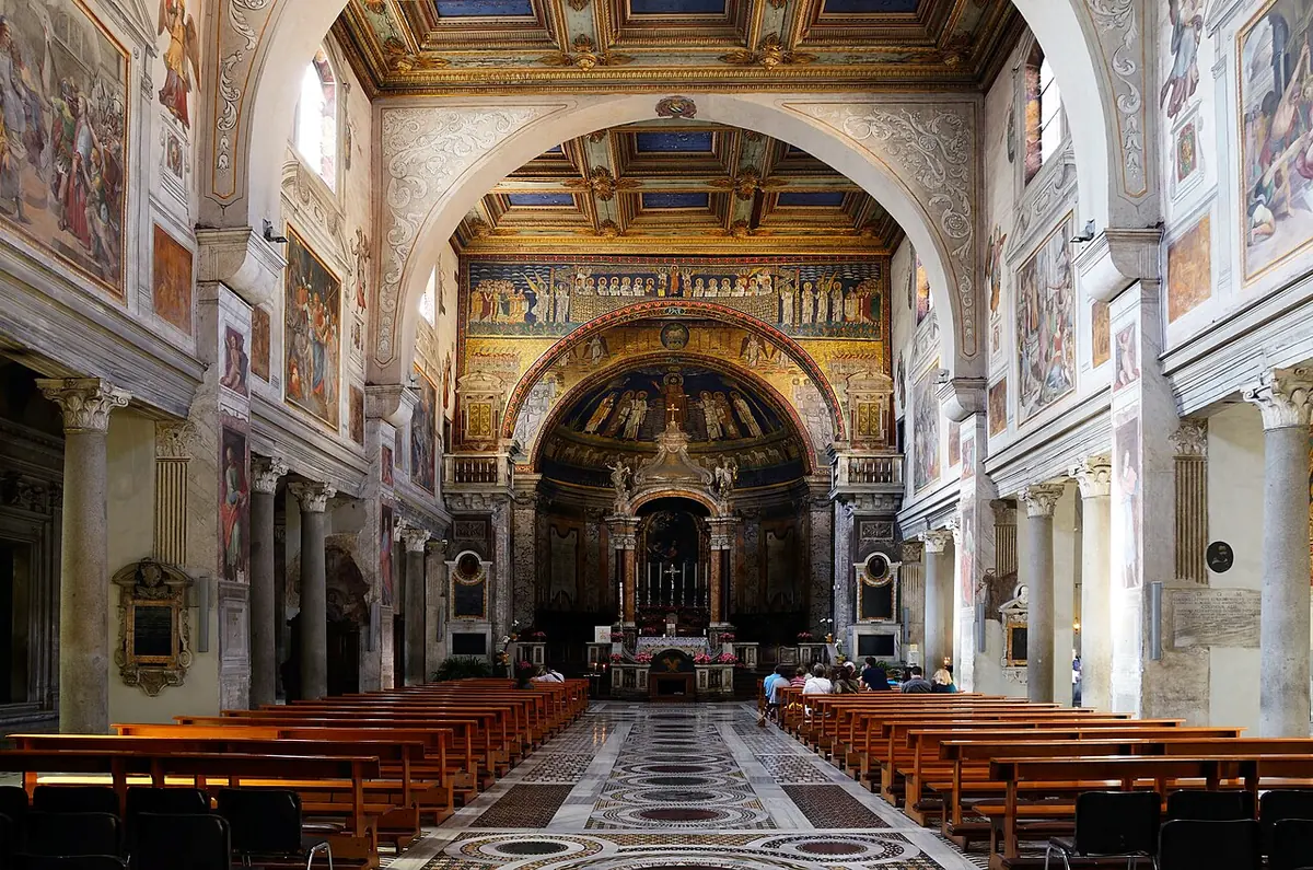 The long nave of a Roman basilica with wooden pews and painted walls, leading to a golden mosaic apse