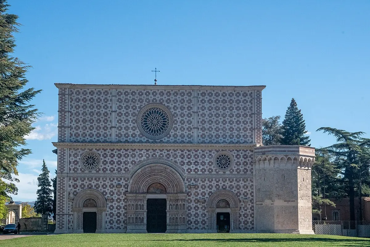 The wide facade of Santa Maria di Collemaggio, patterned in pink and white stone with three round doorways and a rose window, across a green lawn