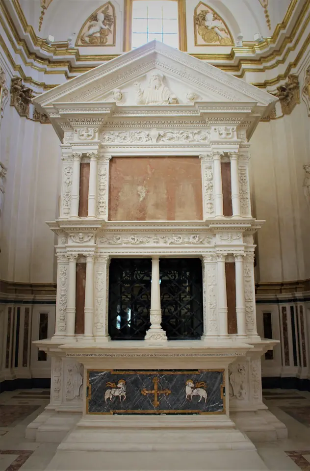 The white Renaissance marble mausoleum of Celestine V in the restored Collemaggio basilica, with columns, a carved frieze and a grille in its center