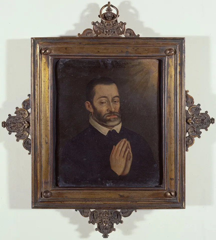 A small dark oil portrait of a bearded man in a black cassock and white collar with his hands joined in prayer, in an ornate gilt frame