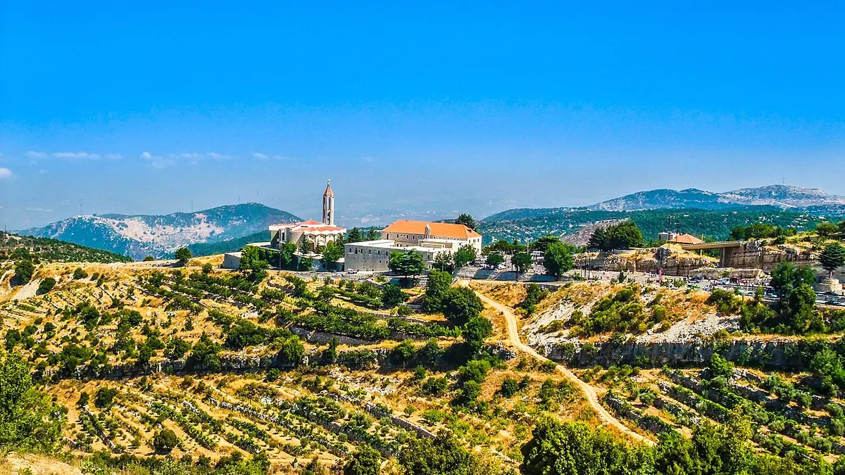 Wide view of a stone monastery with a red-tiled roof and a slender bell tower on a ridge above terraced green hills, with mountains on the horizon