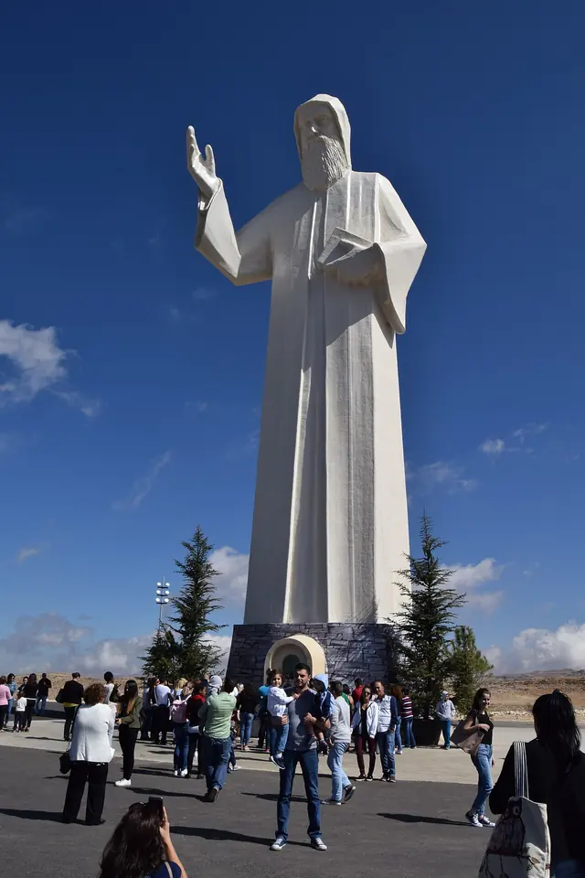 Huge white statue of St. Charbel with one hand raised in blessing on a tall base, with visitors gathered on the plaza below