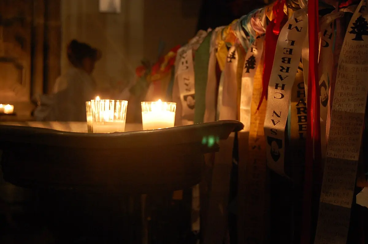 Hundreds of colored ribbons with handwritten prayers hanging beside a row of lit candles in a dark church