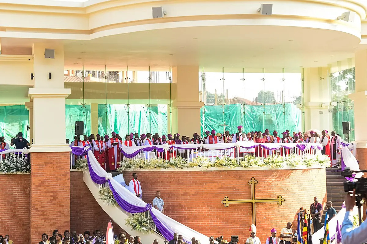 Anglican bishops in red and white robes gathered on a curved raised platform decorated with purple and white cloth before a large crowd