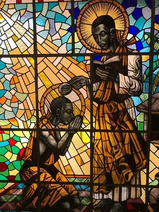 Stained glass of a haloed young man in a long brown robe pouring water from a vessel over the head of a kneeling boy with folded hands