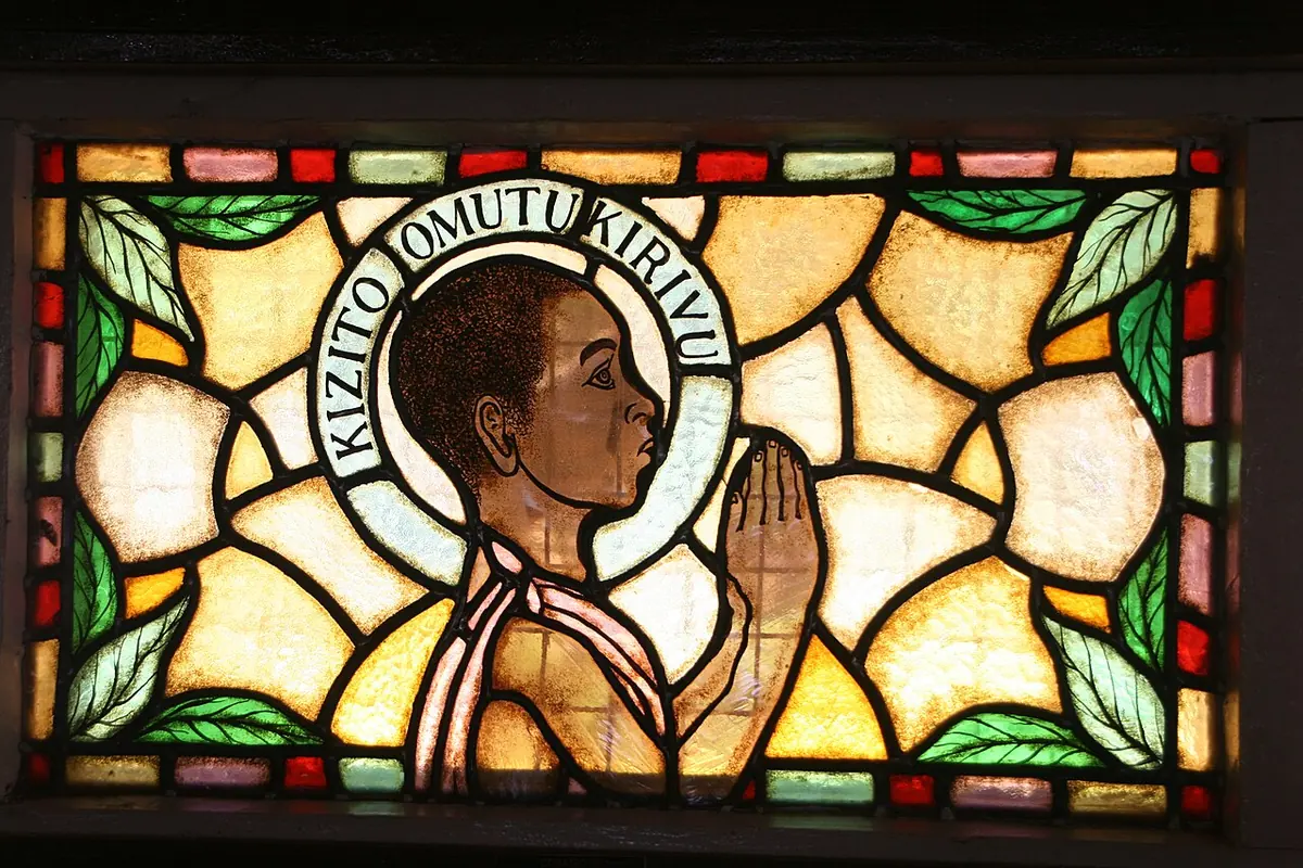 Stained glass of a boy in profile with hands joined in prayer and a halo reading "Kizito Omutukirivu," among golden and green leaves