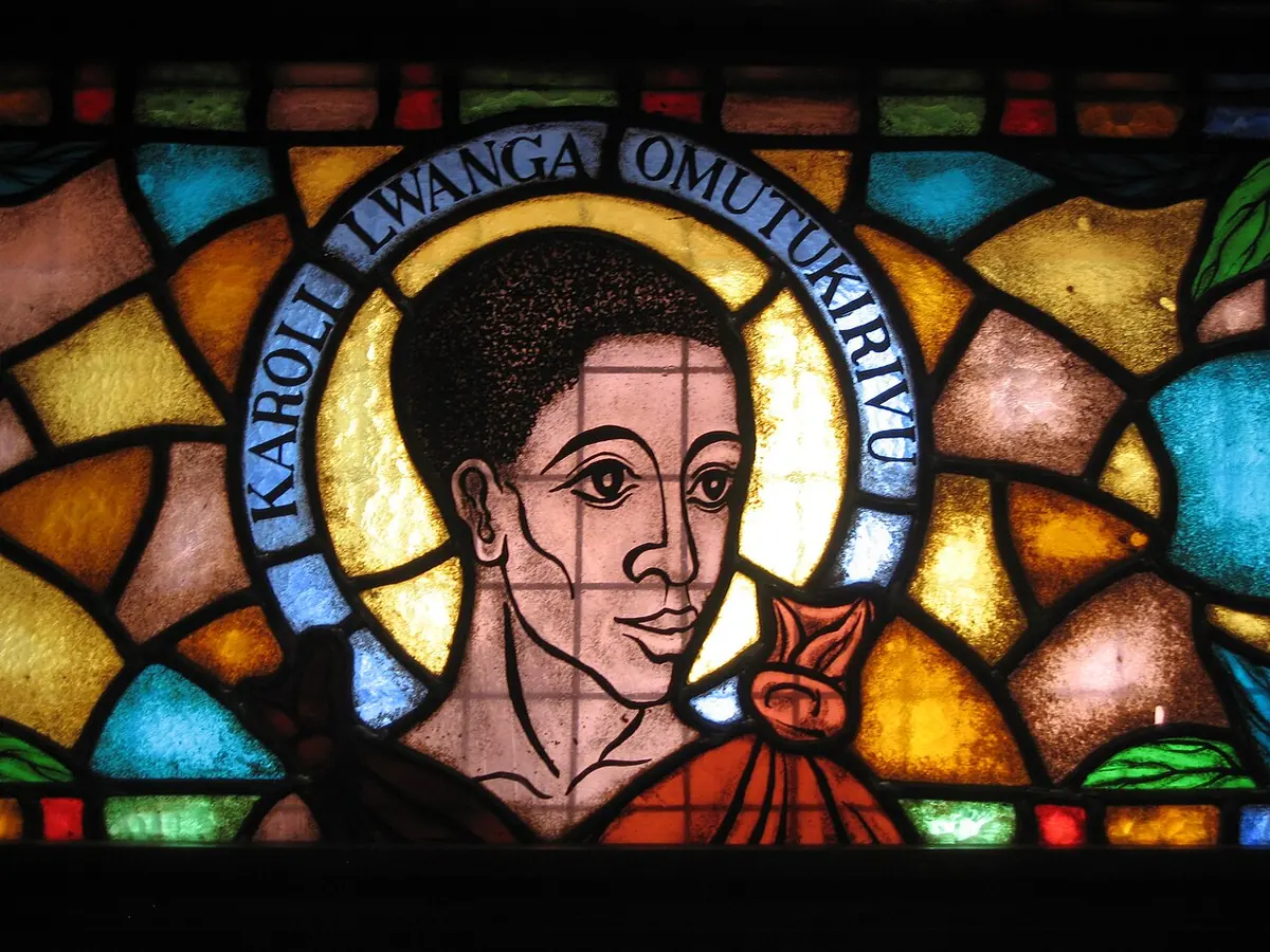 Stained glass of a young man's face with a halo reading "Karoli Lwanga Omutukirivu," set among bright orange, blue and green panes
