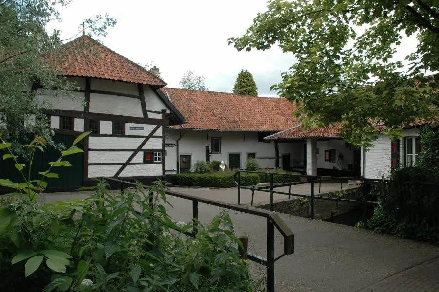 Low whitewashed farm buildings with red tile roofs around a courtyard with trimmed hedges and a small white bust on a pedestal