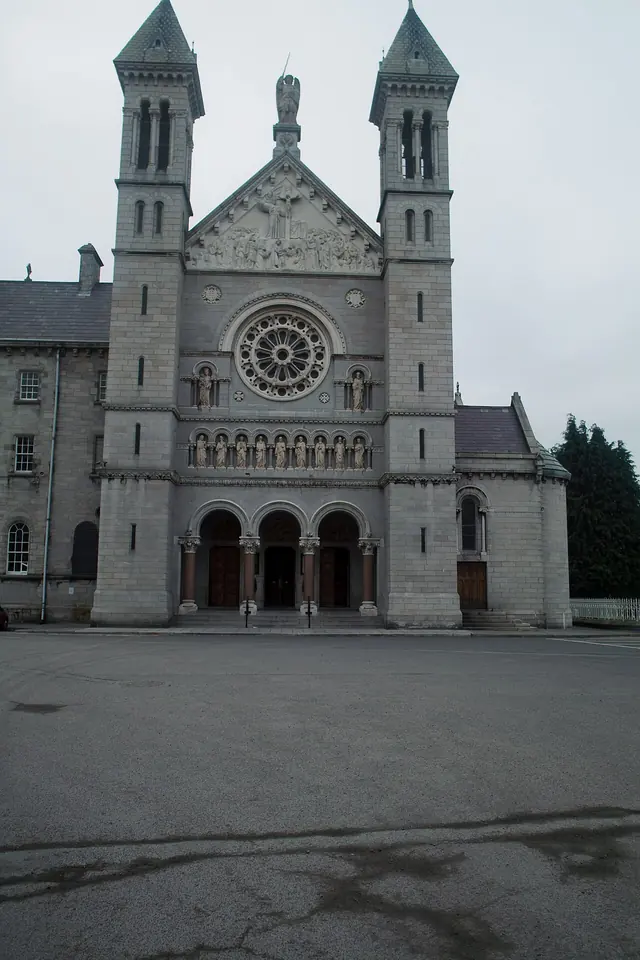 The tall gray stone front of Mount Argus church in Dublin with twin spired towers, a large rose window and three arched doorways