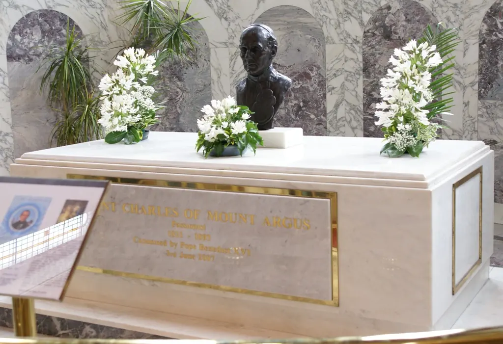 A white marble tomb with a gold inscription panel, a dark bronze bust of the priest on top, and vases of white flowers