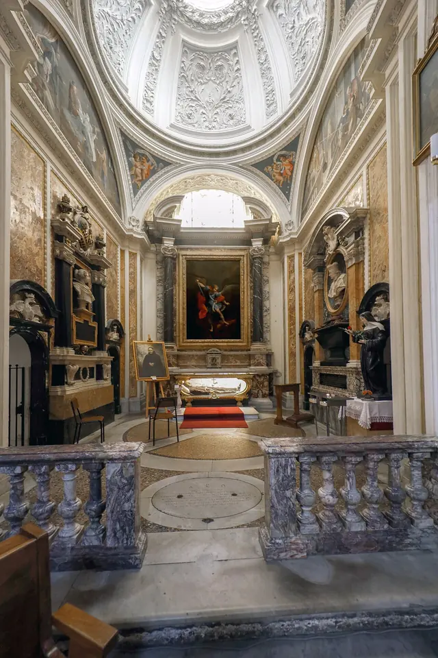 Domed side chapel with white stucco, marble monuments with portrait busts on both walls and an altar with a painting of St. Michael, behind a marble balustrade