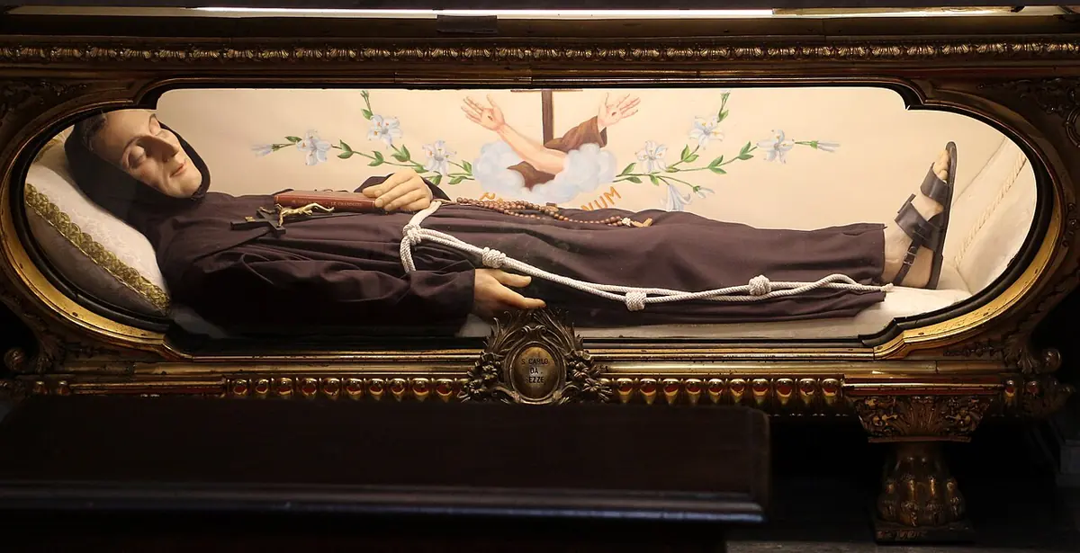 A figure of a Franciscan friar in a brown habit lying with his head on a pillow inside a glass-fronted urn with a gilded frame