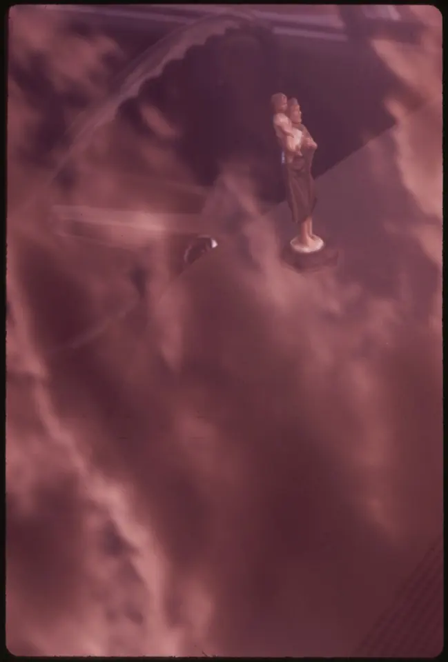 Photograph through a car windshield of a small St. Christopher statuette on the dashboard, with pink clouds reflected in the glass around it