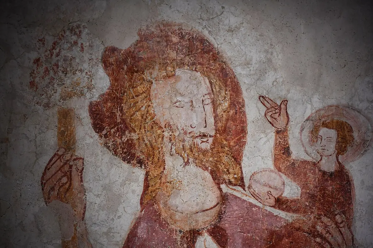 Close-up of a red-outlined medieval wall painting of Christopher's bearded face and his hand holding a staff, with the haloed Christ Child blessing from his shoulder