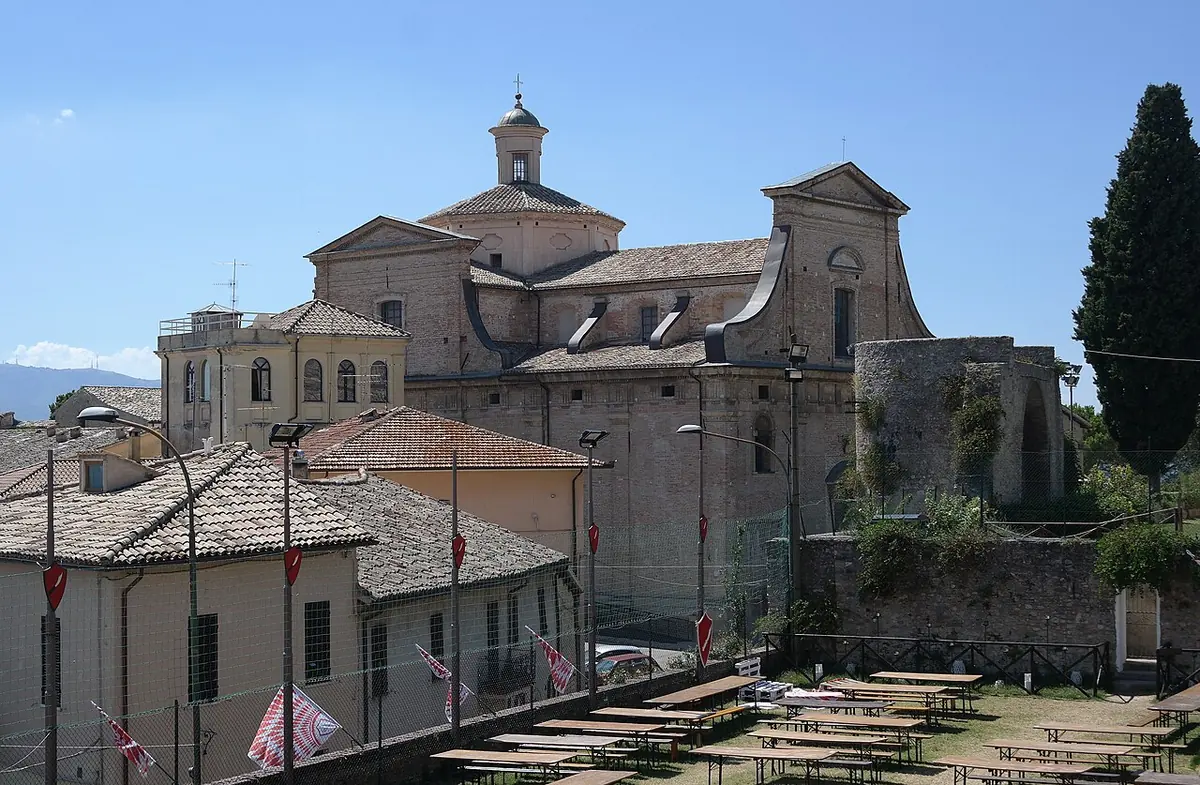 The tan stone church of Santa Chiara in Montefalco with its dome and bell cupola, rising behind tiled rooftops and a lawn on a sunny day