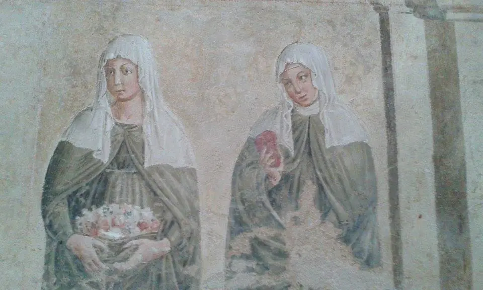 A faded fresco of two women saints in pale veils side by side, one holding an apron full of roses and the other holding a red heart