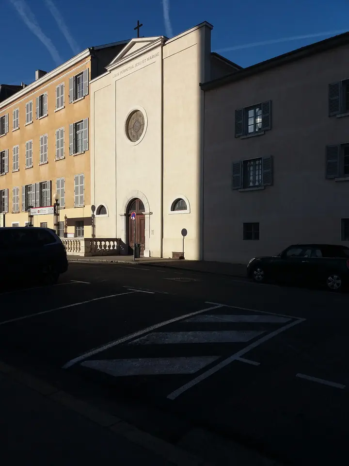 A plain classical chapel facade with a round window and a cross, between tall apartment buildings on a square