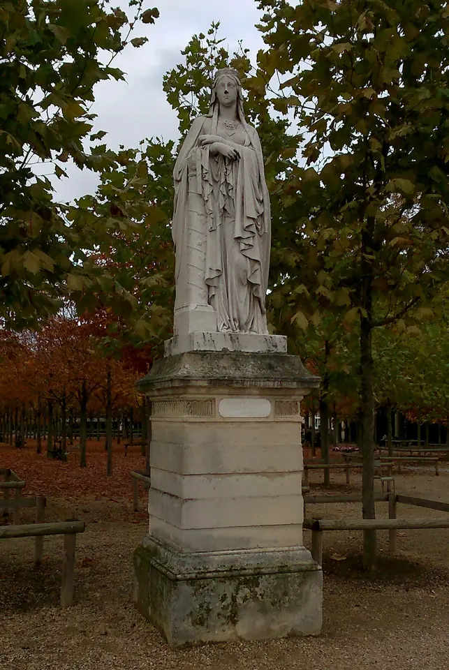 White marble statue of St. Clotilde as a veiled queen with joined hands, standing on a tall pedestal under autumn trees