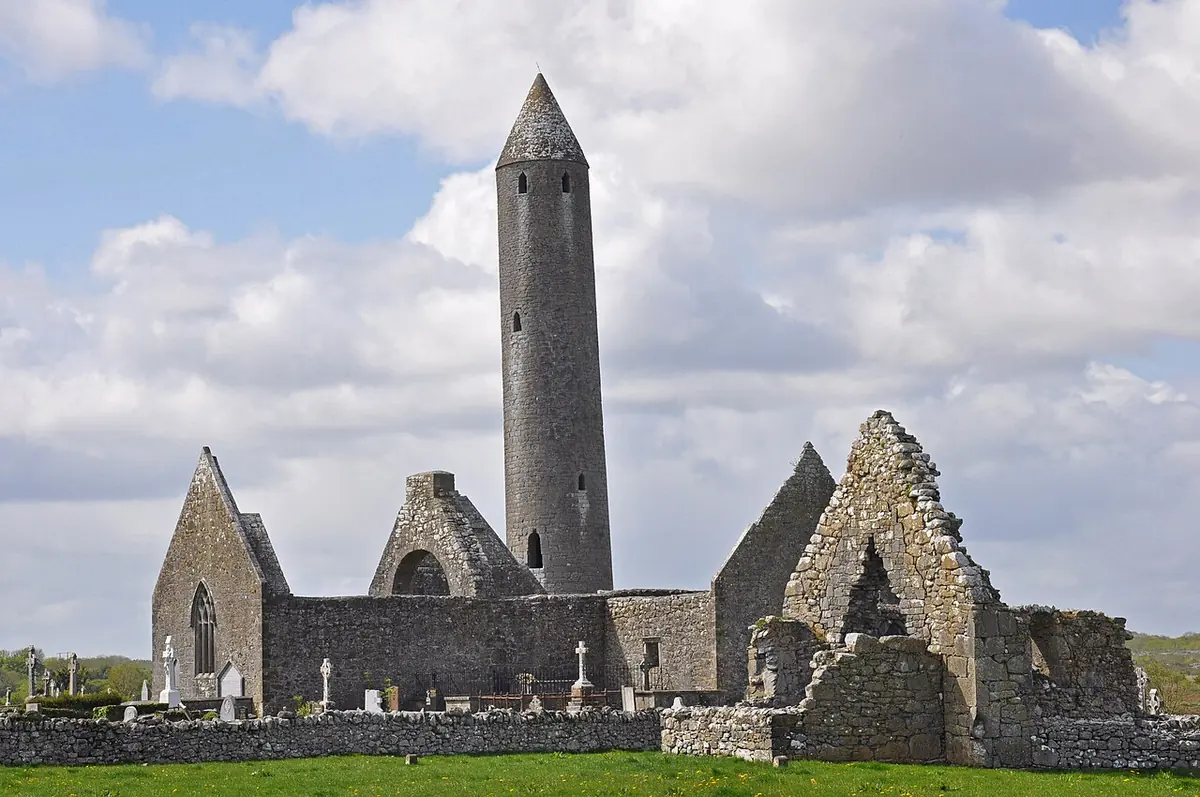 A tall stone round tower that leans slightly, rising among the roofless gray stone churches and graves of an old monastic site under a cloudy blue sky