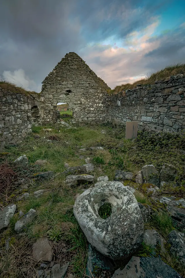 The roofless gable of a small old stone church on Inishbofin under a dramatic evening sky, with a holed stone among the grass and rocks in front