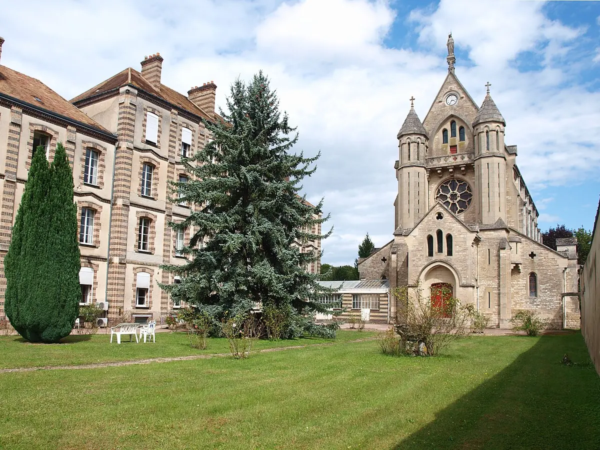 A neo-Gothic stone chapel with a rose window and slender towers beside a long convent building and evergreen trees on a lawn