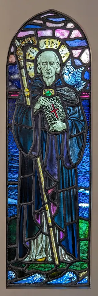Stained-glass figure of St. Columba as a gray-haired monk in a blue cloak with a halo, holding a staff and a red book, standing above the sea