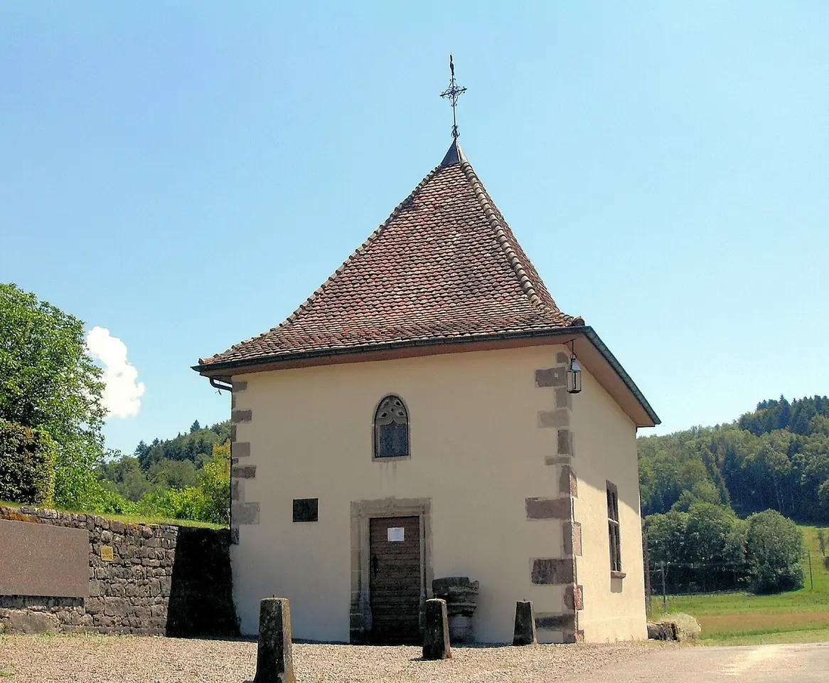 A small cream-colored chapel with a steep tiled roof and a cross on top, standing in a meadow at the edge of a wooded valley
