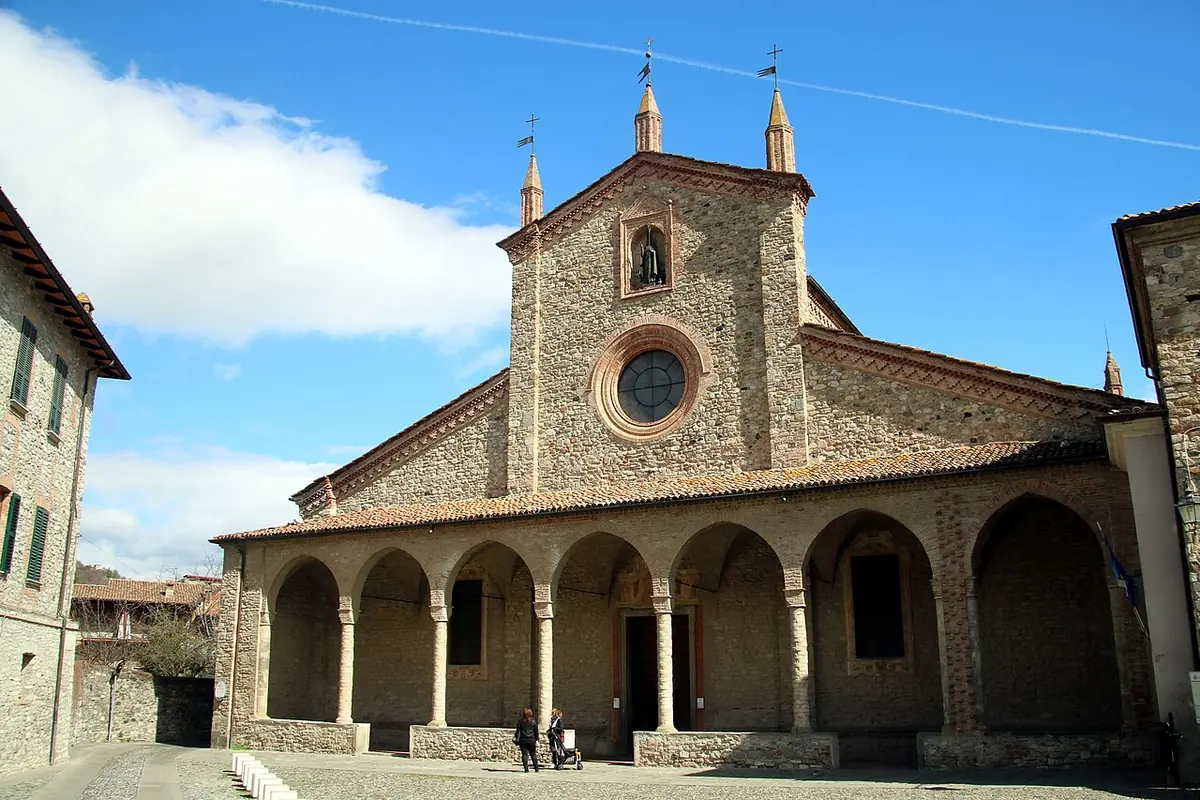 The brick facade of the abbey church at Bobbio with a rose window, small spires and an arched portico, on a cobbled square under a blue sky