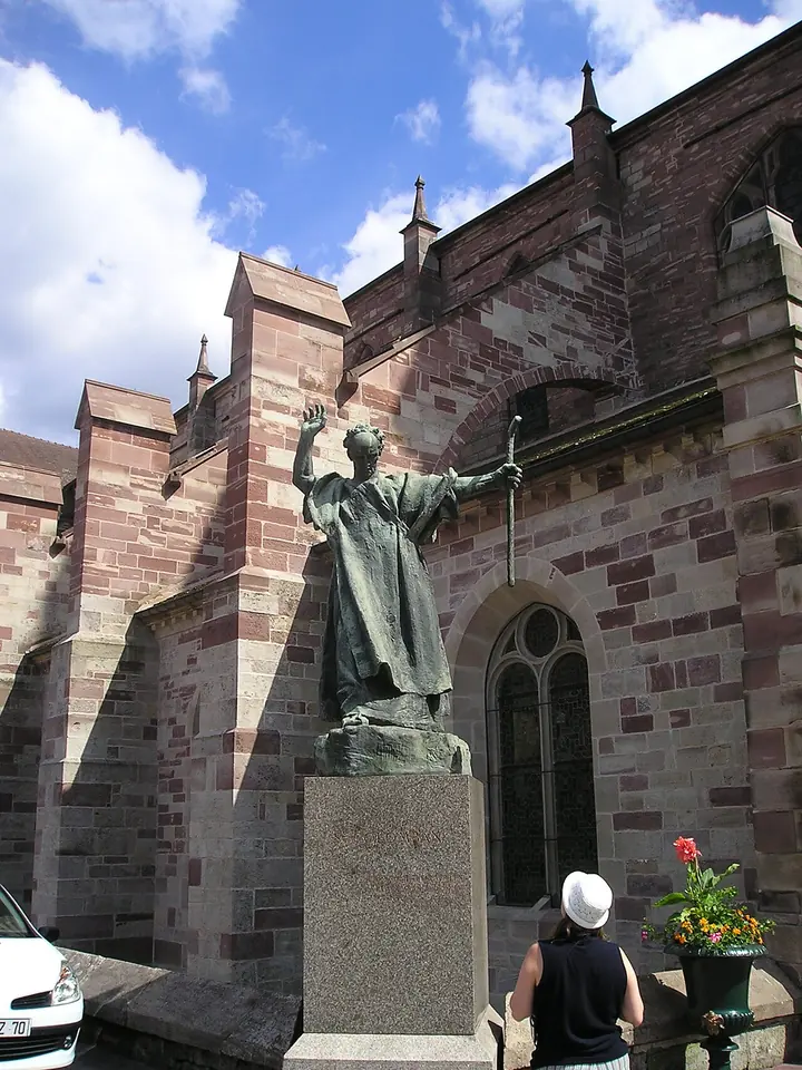 Bronze statue of a monk with one arm raised high and the other holding a staff, standing on a stone pedestal beside the red sandstone wall of a Gothic church