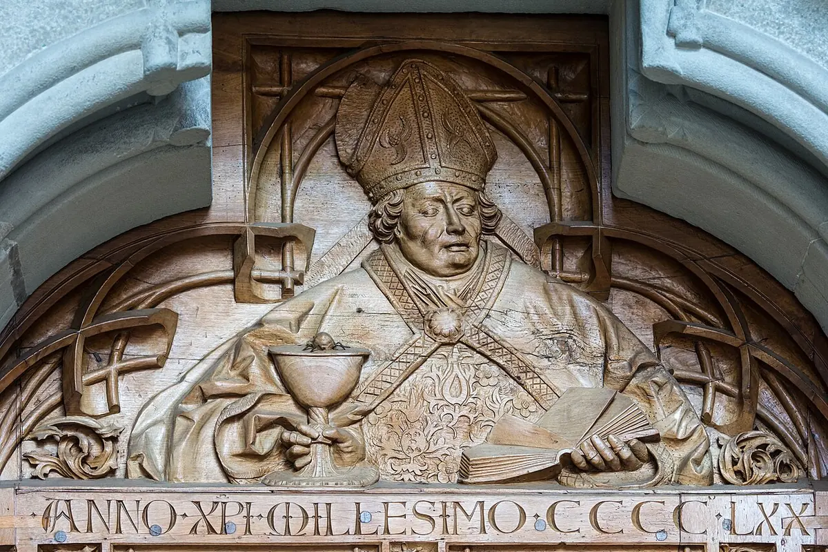 Carved wooden relief of a bishop in a miter with a chalice before him, above a Latin inscription giving the year 1470