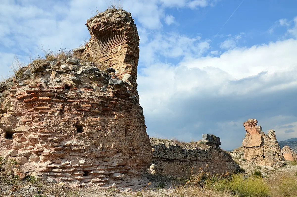 Weathered towers and walls of old brick and stone rising against a blue sky with white clouds, with dry grass in front