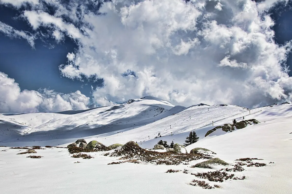Snow-covered rounded mountain slopes with scattered rocks and a few dark fir trees under a dramatic sky of white clouds