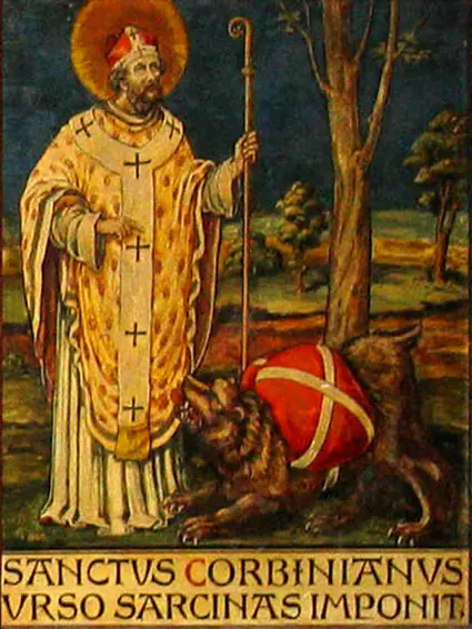 Painted panel of a bishop in a golden chasuble with crosses and a red miter, holding a crosier, as a brown bear carrying a red pack crouches at his feet, with a Latin caption below