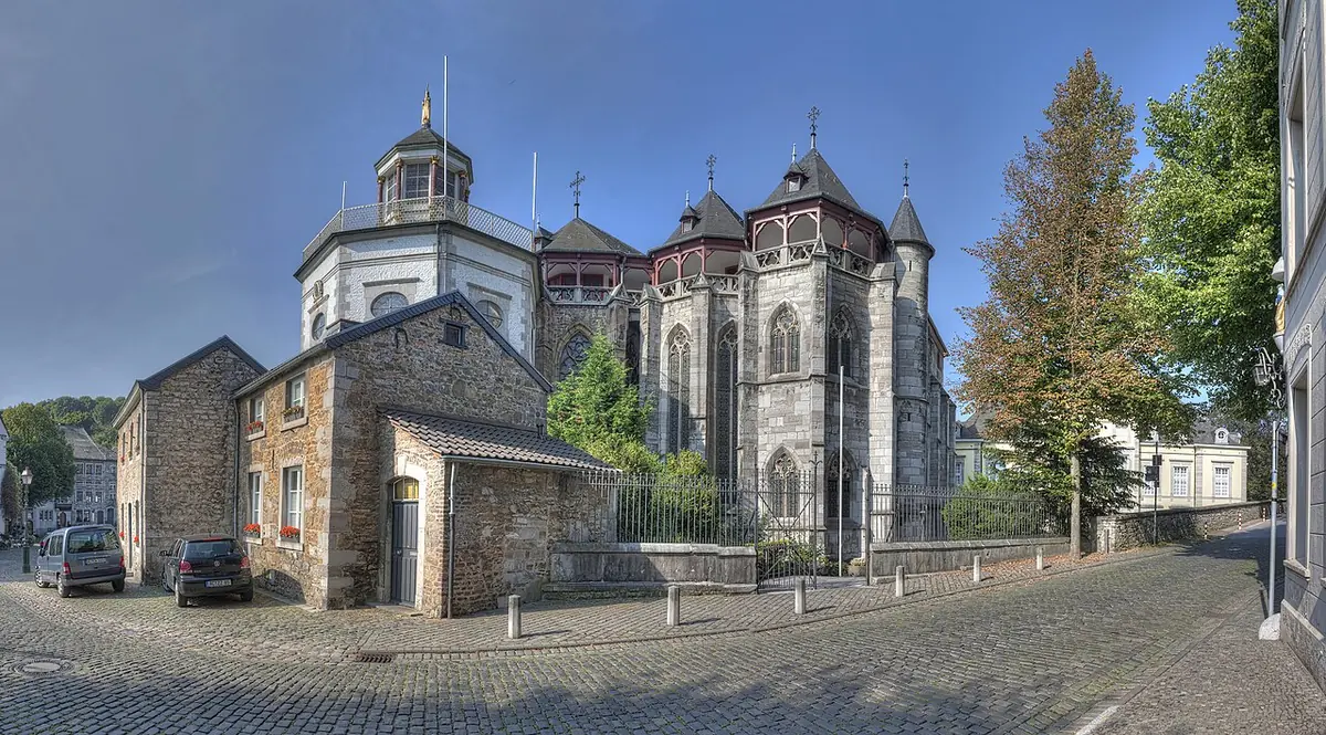 Wide view of a large gray stone Gothic church with several pointed roofs and an octagonal domed chapel at its east end, beside a cobbled square