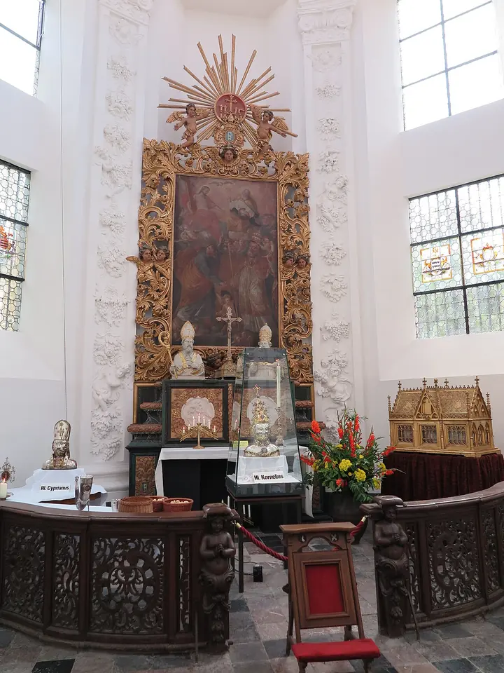 A bright white Baroque chapel with a gilded altarpiece and sunburst, and silver reliquaries and a golden shrine set out before the altar behind a carved wooden rail