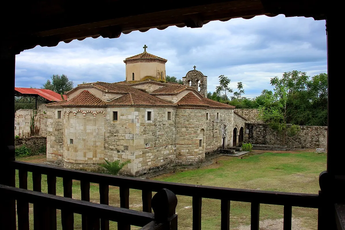 A stone church with a tiled dome in a grassy monastery courtyard, framed by the dark wooden railing of a porch