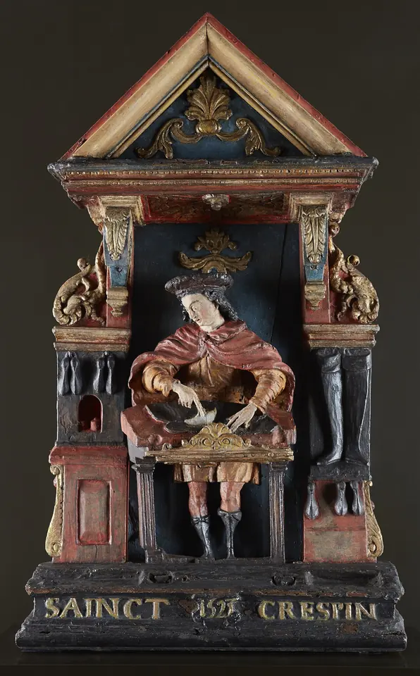 Painted wooden shop sign shaped like a small shrine, with a carved St. Crispin cutting leather at his bench beside a pair of tall boots, inscribed "Sainct Crespin"