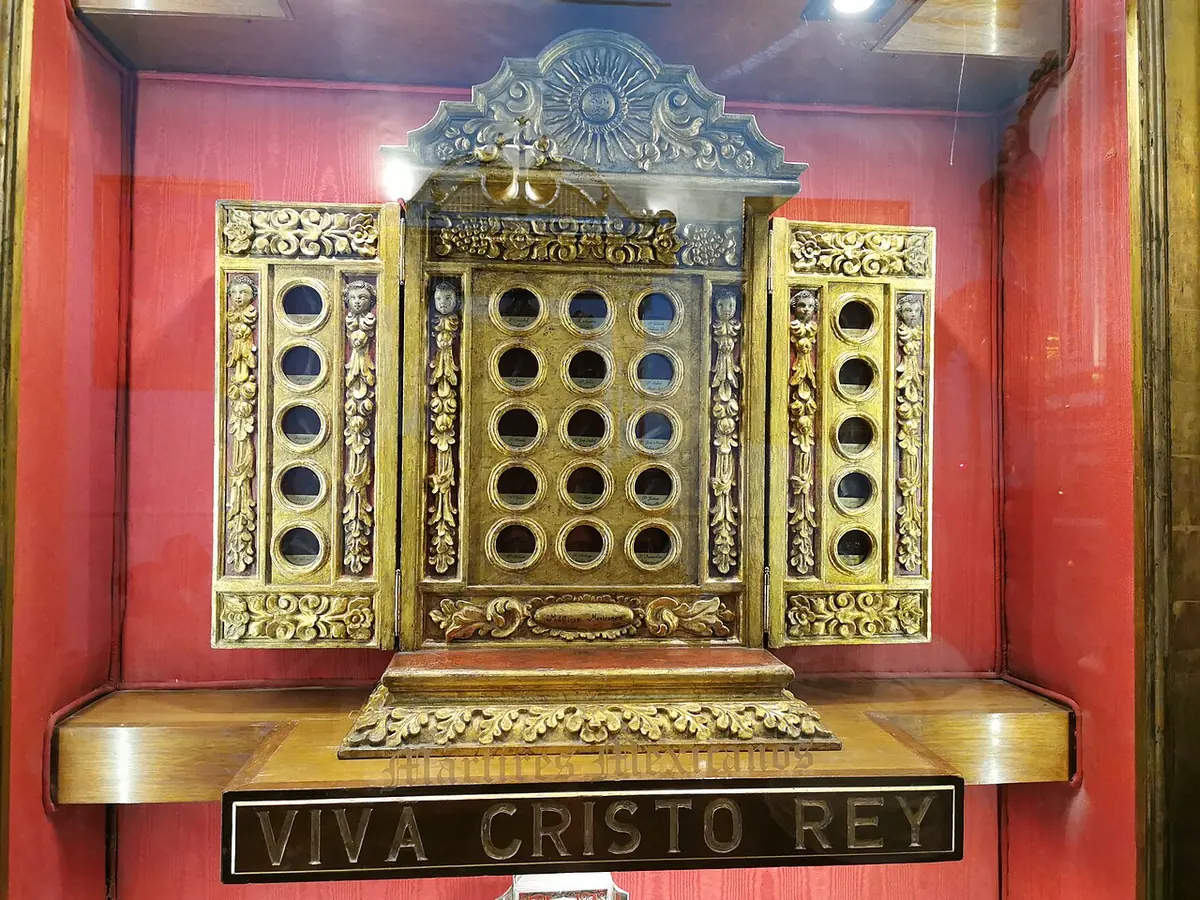 A gilded wooden reliquary with rows of small round windows, displayed on a red silk background above a plaque reading Viva Cristo Rey