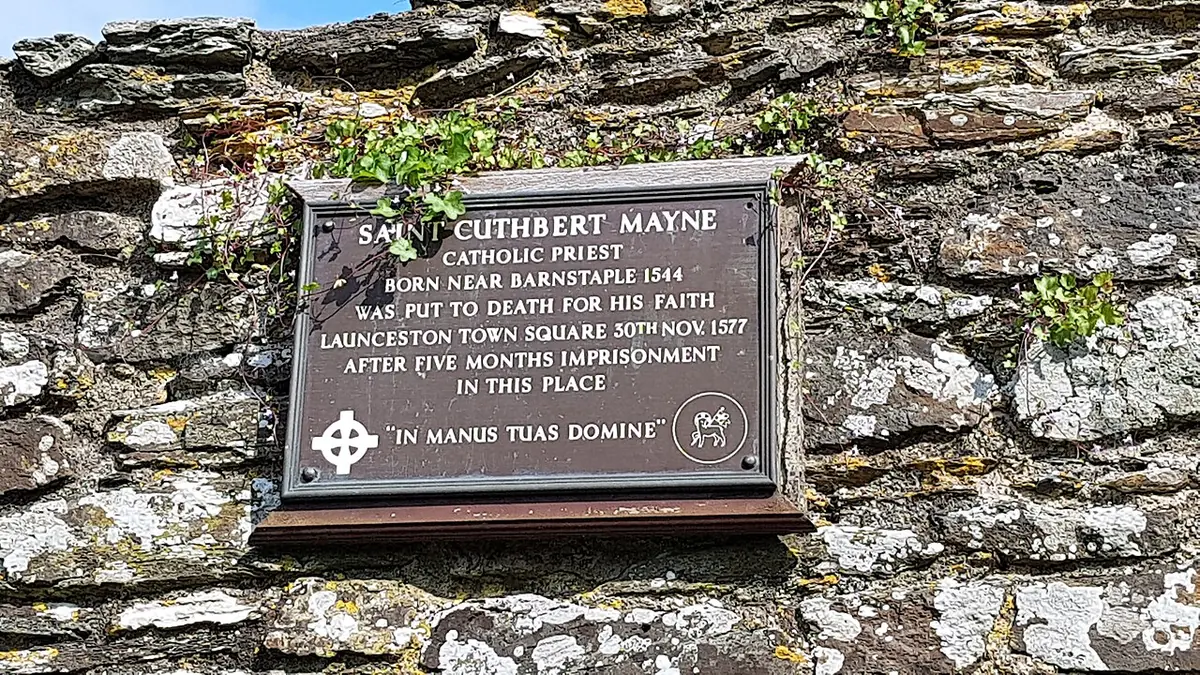 A dark bronze plaque on an old stone wall recording that St. Cuthbert Mayne, Catholic priest, was put to death for his faith in Launceston town square, ending with the words In manus tuas Domine