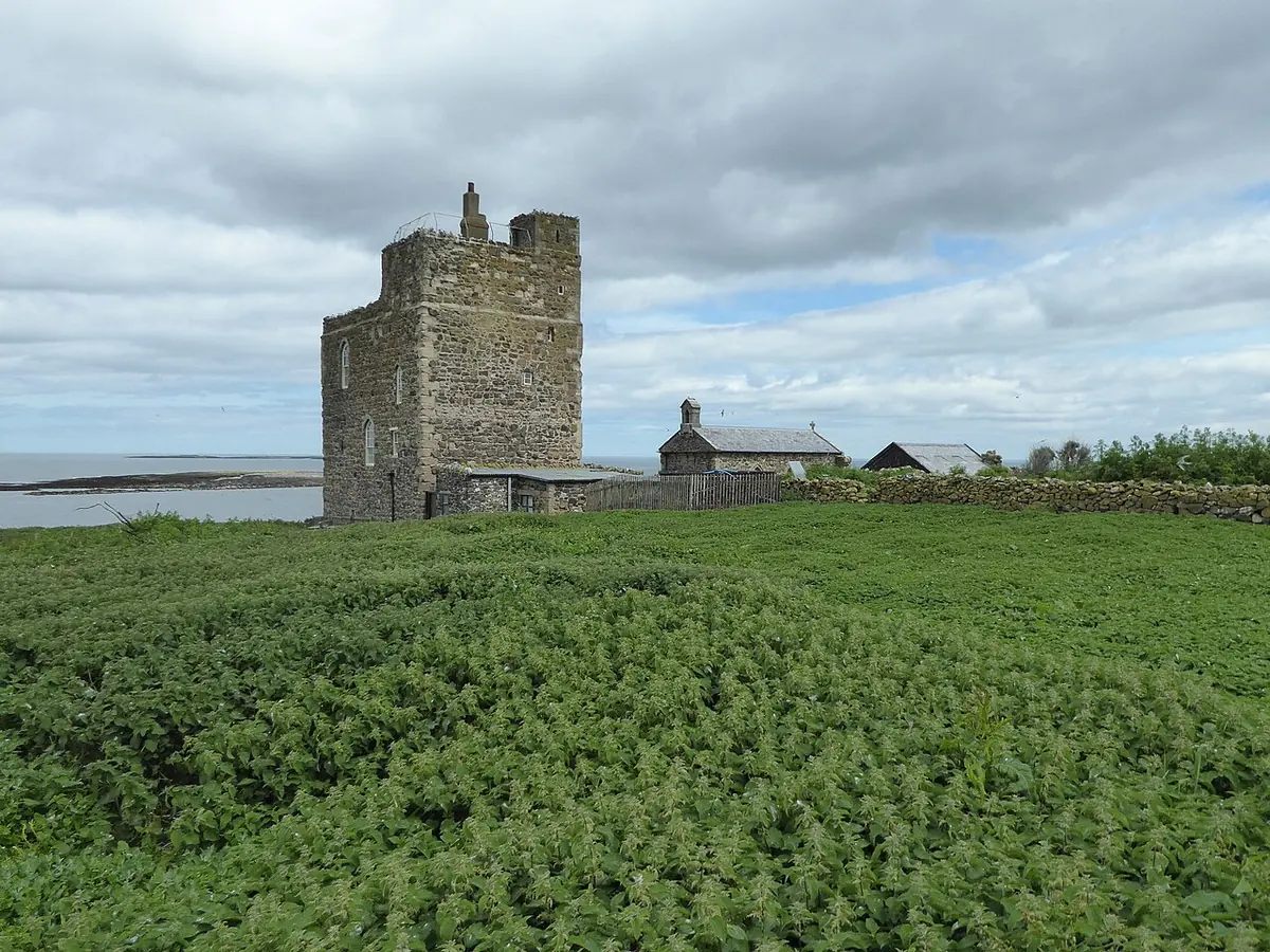 A square stone tower and a small stone chapel on a low green island under a cloudy sky, with the sea beyond