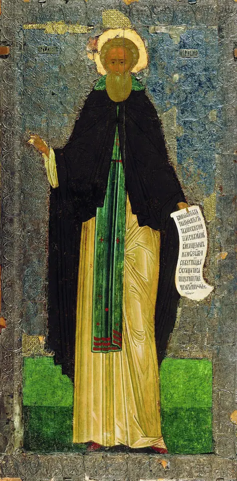 A tall, narrow icon of a monk with a halo and long beard, in a dark mantle over a golden robe, holding an unrolled white scroll and raising his right hand