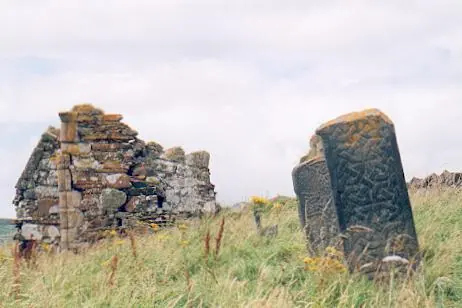 A crumbling stone church wall and a weathered carved cross slab standing in long grass on an island